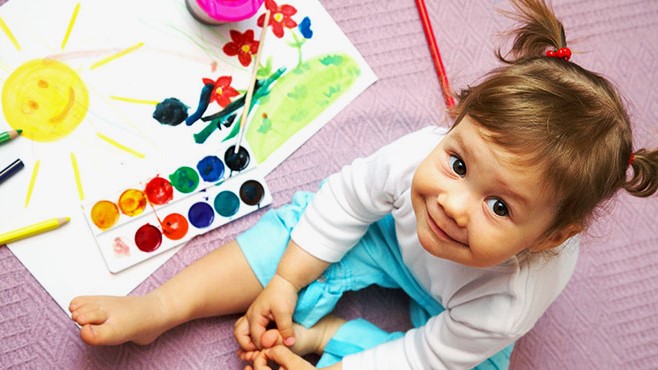 A young girl is sitting on the ground with a water color paint set and a partially completed painting of flowers growing under a yellow sun.