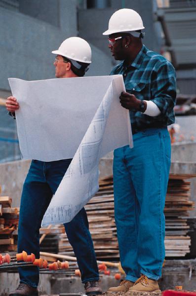 Two men are examining blueprints on a construction site. They are both wearing the same white hardhat and looking in the same direction; to their right.