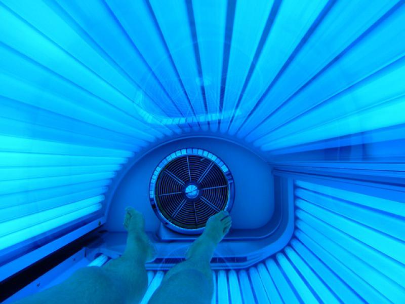 A person laying in a tanning bed. You can see their legs extending from out of frame towards the bottom of the tanning bed. There is a large fan directly beneath their feet. The tanning bed is filled with cylindrical lights that have a light blue glow.