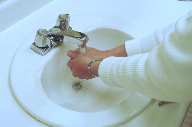 A person is seen washing their hands in a white sink with a chrome faucet. They have a white shirt on and a silver bracelet on their left arm.