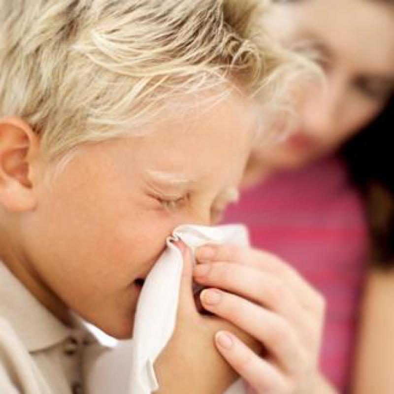 A child is seen blowing their nose into a small white tissue. Their parent is assisting them by helping hold the tissue in place.