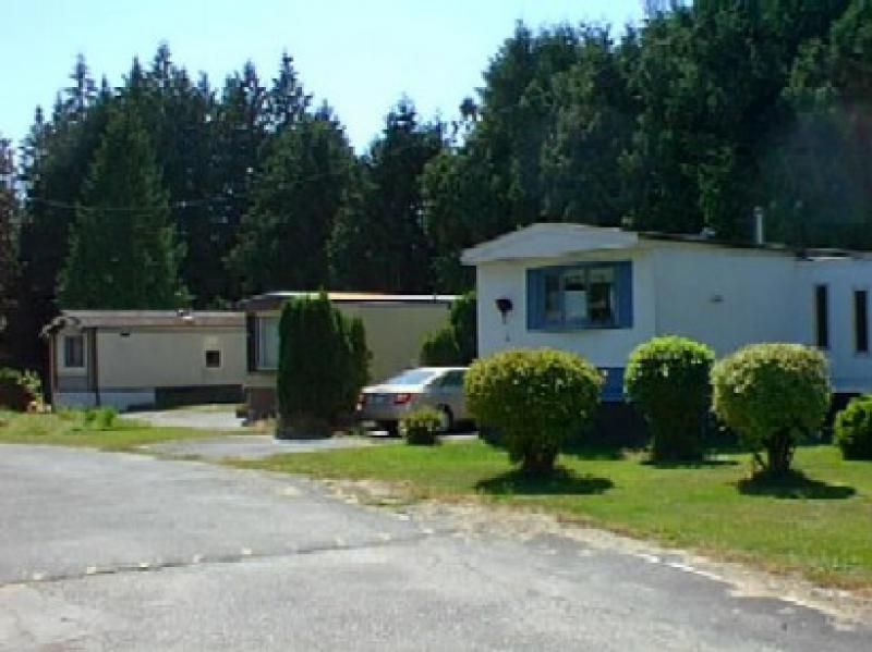 A side view of three mobile homes. There is a sedan in the driveway of the frontmost mobile home next to a hedge line. There are some small, well-pruned bushes in front of the frontmost mobile home.