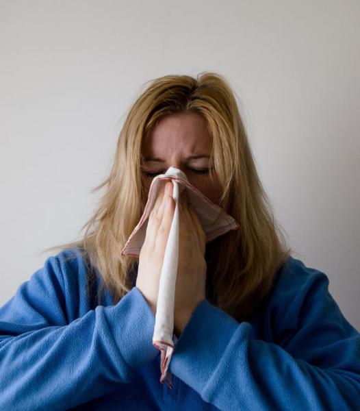 A person is seen coughing into a cloth. They are fully covering their nose and mouth with the cloth. They are wearing a blue sweater and have long dirty-blond hair.