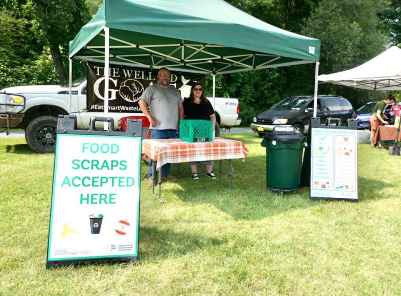 A  stand at a local event  with signs stating "Food scraps accepted here." They are under a green tent with a large waste bin and a table under the tent.
