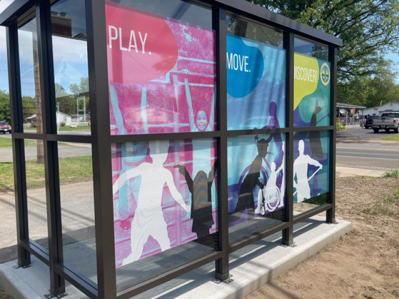 A bus stop with three decorative posters on the back of it. The posters say "Play." "Move." and "Discover!"