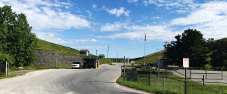 Clinton County Landfill enterance road, chainlink fencing and gate.  Trees and hill with blue sky and clouds.